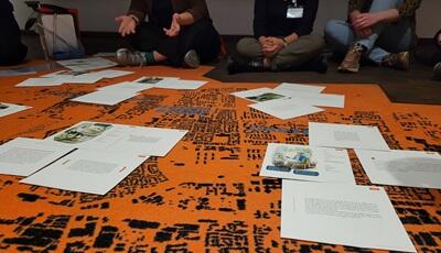 Mehrere Personen sitzen auf einem orangen-schwarzen Teppich. Auf dem Teppich liegen Impulskarten mit Zitaten und Abbildungen.