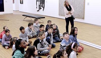Schüler*innen sitzen im Germanischen Nationalmuseum auf dem Boden und betrachten ein Exponat, im Hintergrund steht die Lehrerin
