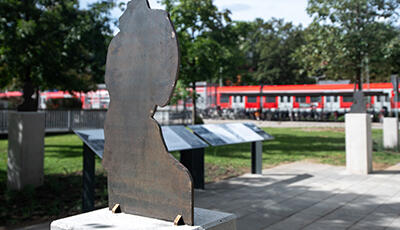 Erinnerungsort Flachsröste Lohhof: Grünanlage, im Vordergrund ist eine Skulptur sichtbar, im Hintergrund Texttafel und noch weiter weg ein roter Zug.