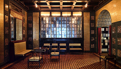 Prächtiger Raum mit einer Spiegelwand, goldenen Wandverzierungen, einer Kassettendecke, einigen Stühlen und einem Tisch. Rechts ist ein bogenförmiger Durchgang in den nächsten Raum erkennbar.