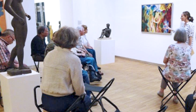 Eine Seniorengruppe sitzt auf Klappstühlen im Museum vor einem Kunstwerk.