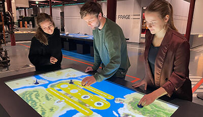 Medientisch in einem Ausstellungsraum des tims, an dem drei Personen arbeiten.