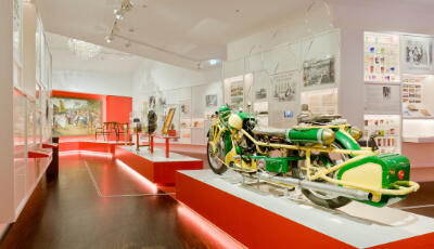 Blick in einen Ausstellungsraum des Sudetendeutschen Museums. Im Vordergrund ist ein altes Motorrad zu sehen, auf dem drei Personen fahren konnten. Im Hintergrund Vitrinen mit farbigen Gläsern.
