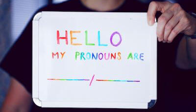 Eine Hand hält ein Schild in die Kamera, auf dem in bunten Buchstaben steht: Hello, my pronouns are --- / ---