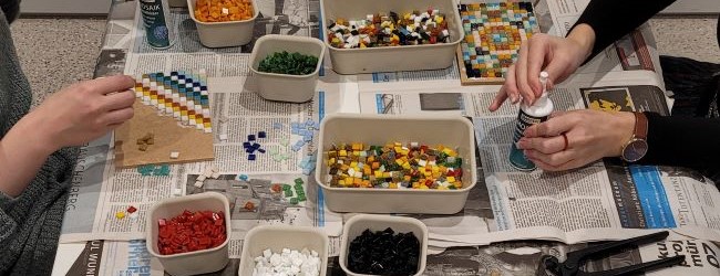 Blick auf einen mit Zeitung ausgelegten Tisch. Zwei Personen kleben bunte Mosaikplatten.